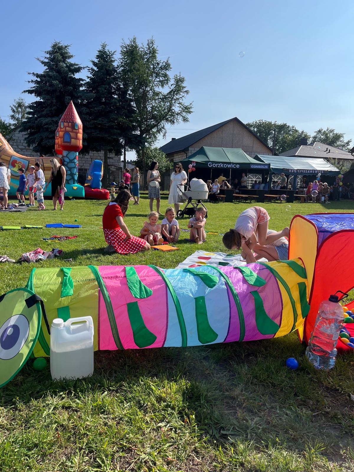 Kolorowy tunel dla dzieci na trawie, w tle dmuchany zamek, grupa dzieci i dorosłych. Plenerowa impreza z atrakcjami dla najmłodszych w słoneczny dzień.
