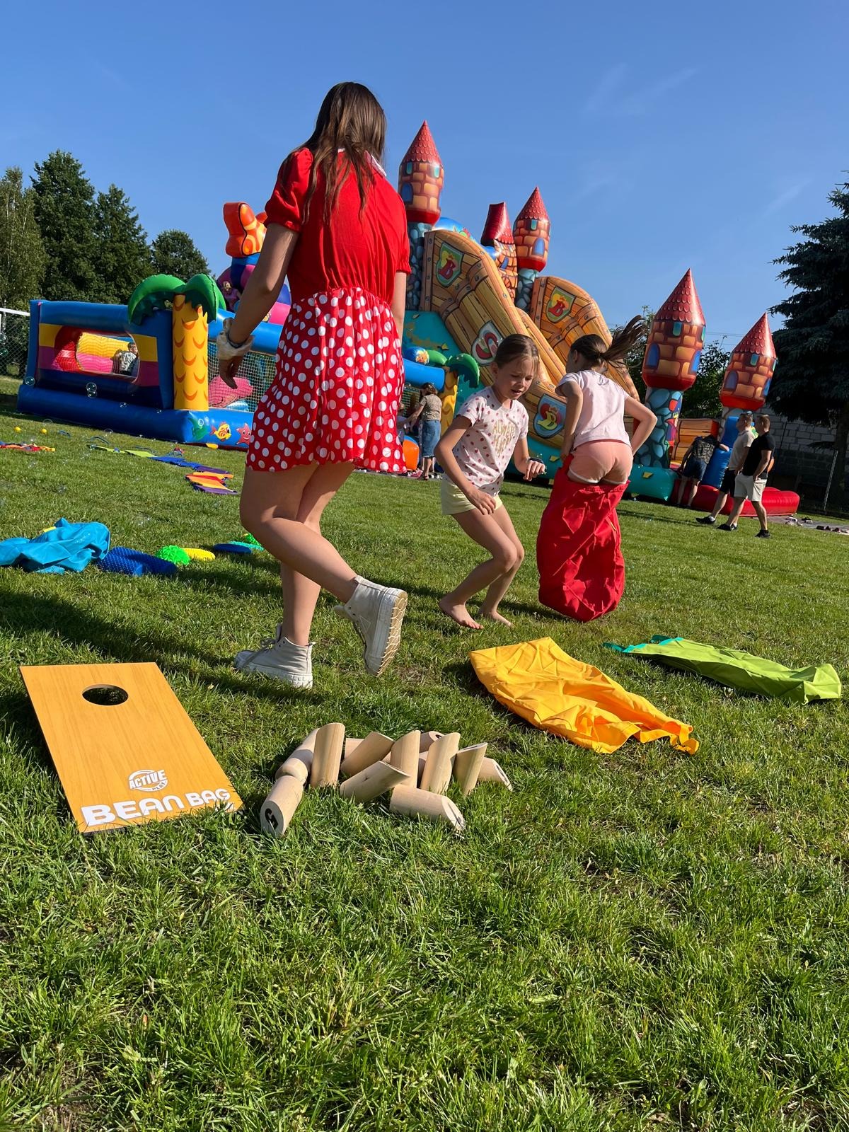 Dzieci bawią się na trawie podczas imprezy plenerowej: skoki w workach, gra w kręgle i cornhole. W tle dmuchany zamek i animator w stroju.
