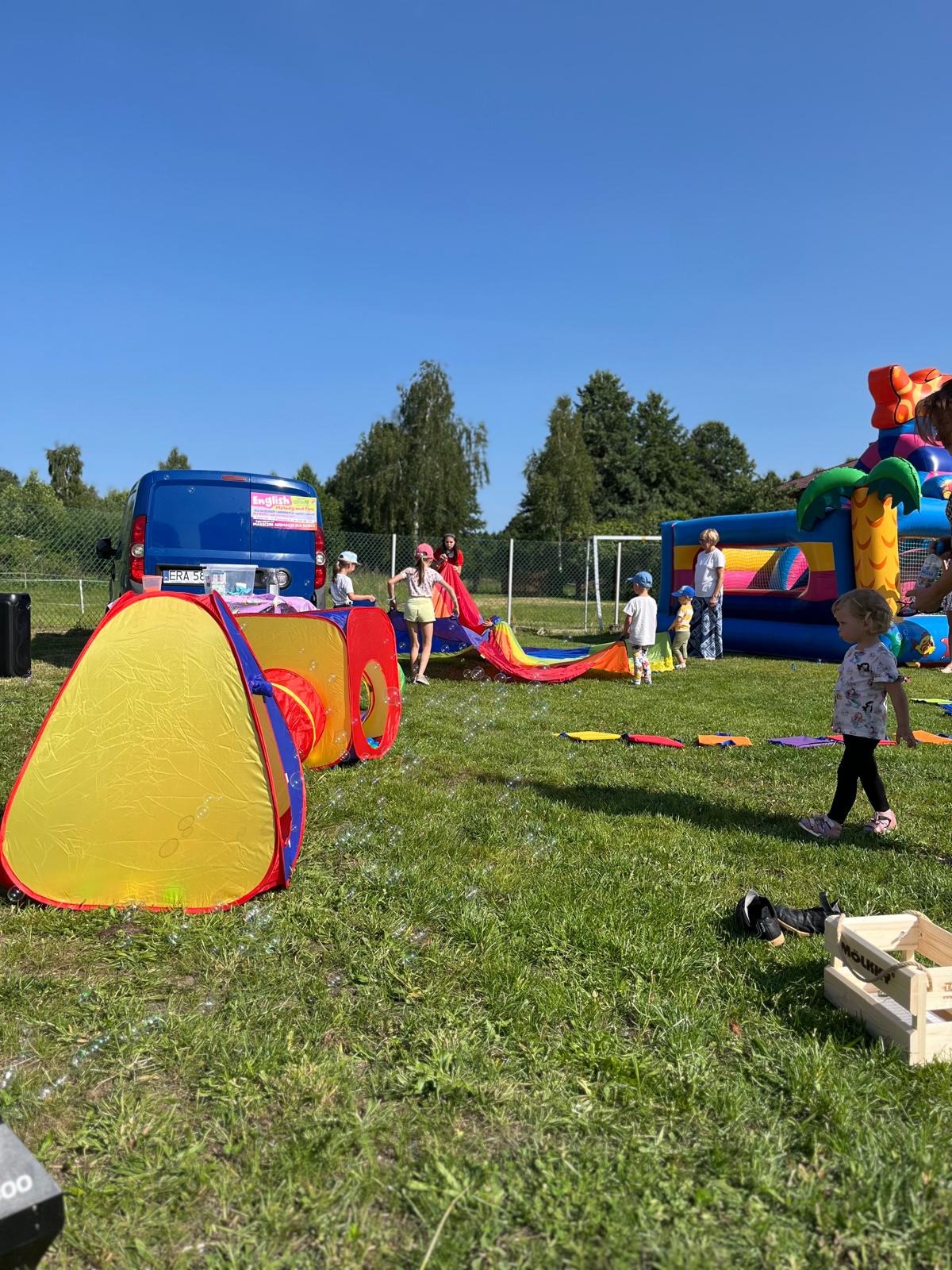Plenerowa impreza dla dzieci: kolorowy dmuchaniec, tunel, chusta animacyjna i dzieci na trawie w słoneczny dzień. W tle niebieski bus.