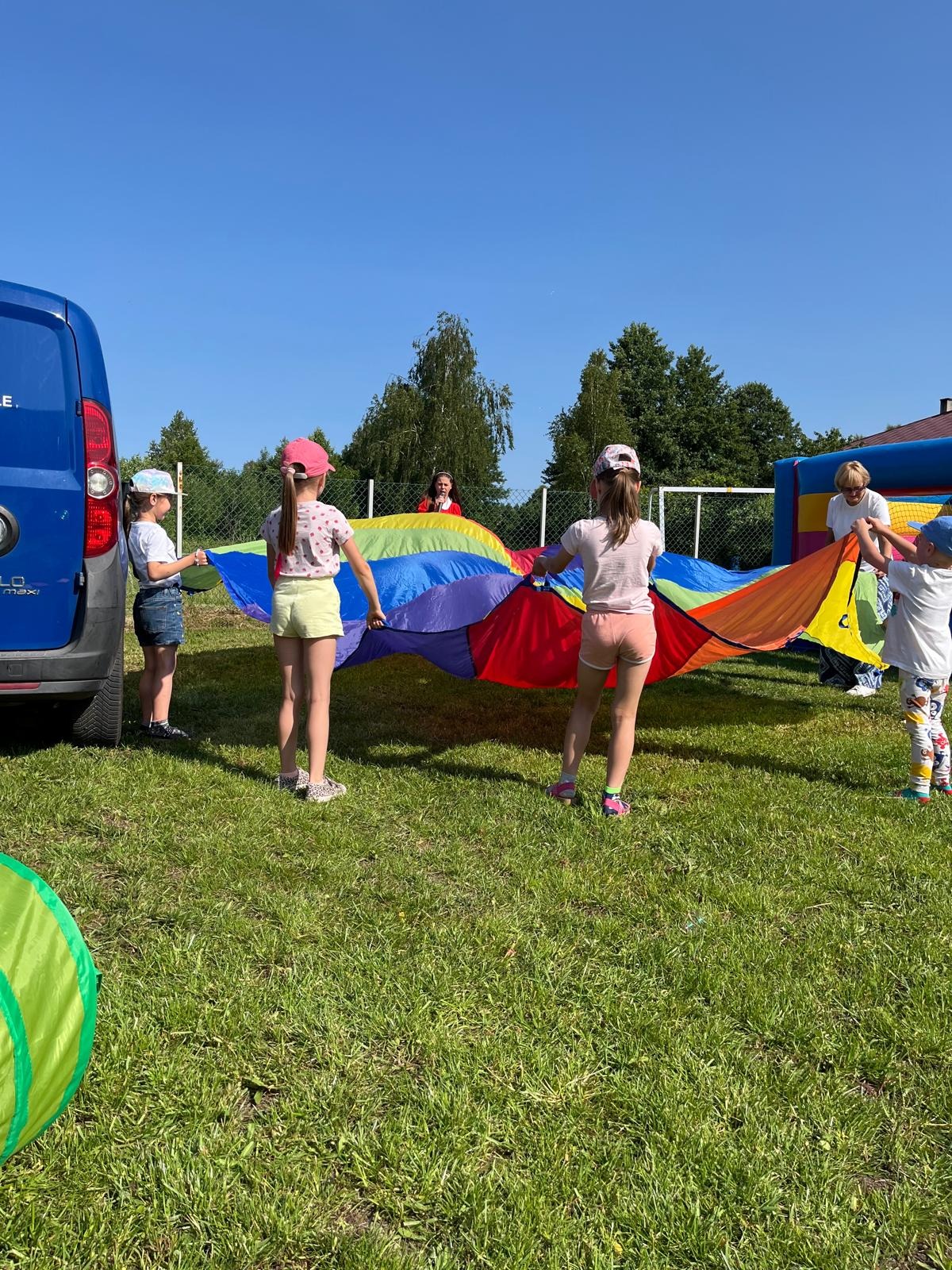 Grupka dzieci bawi się kolorową chustą animacyjną na trawie, w tle częściowo widoczny dmuchany zamek i niebieski van, słoneczny dzień.