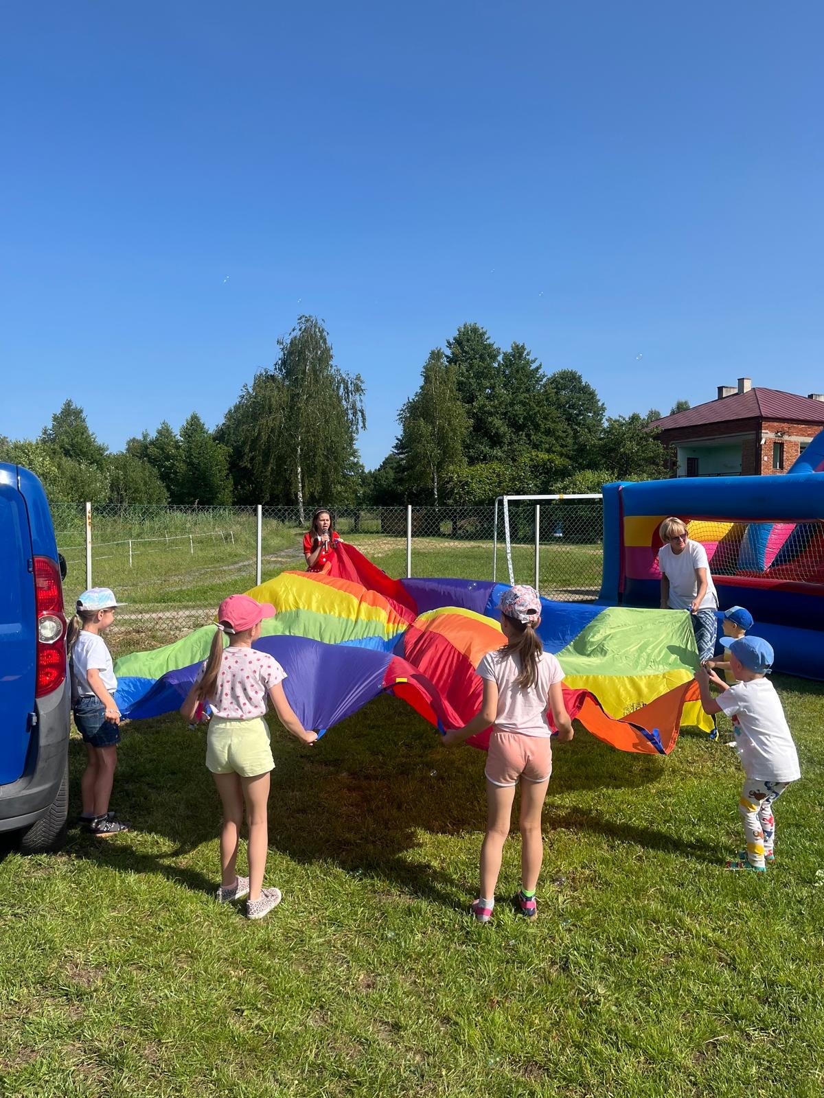 Dzieci bawią się kolorową chustą animacyjną na trawie, w tle dmuchaniec i animator w czerwonej sukience. Słoneczny dzień, błękitne niebo.
