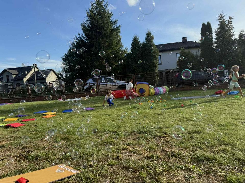 Dzieci bawią się na trawie w otoczeniu baniek mydlanych, kolorowych mat i tunelu. W tle domy i drzewa, słoneczny dzień. Radosna atmosfera imprezy plenerowej.