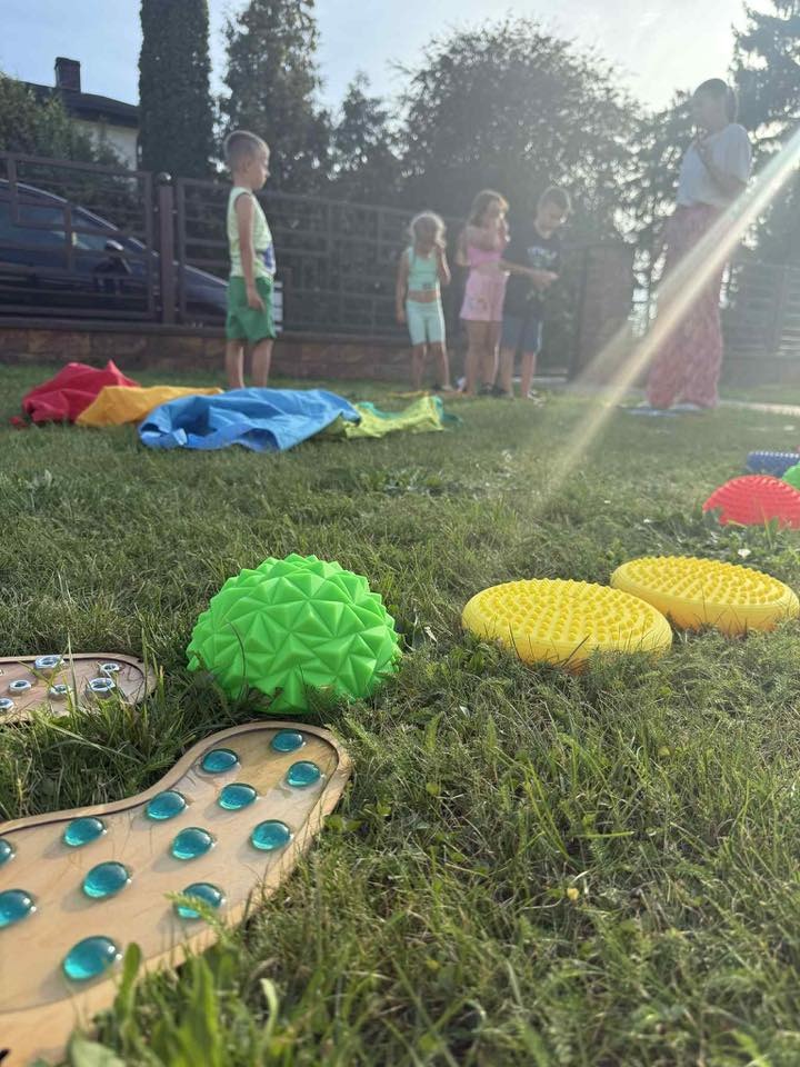 Akcesoria do animacji na trawie: kolorowe poduszki sensoryczne i ścieżka z wypustkami. W tle grupa dzieci i animatorka, częściowo zasłonięci blaskiem słońca.
