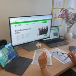 Biuro rachunkowe ATUT: monitor z otwartą stroną, laptop, tablet, dokumenty, kalkulator, okulary. Kobieta przegląda dokumenty na biurku.