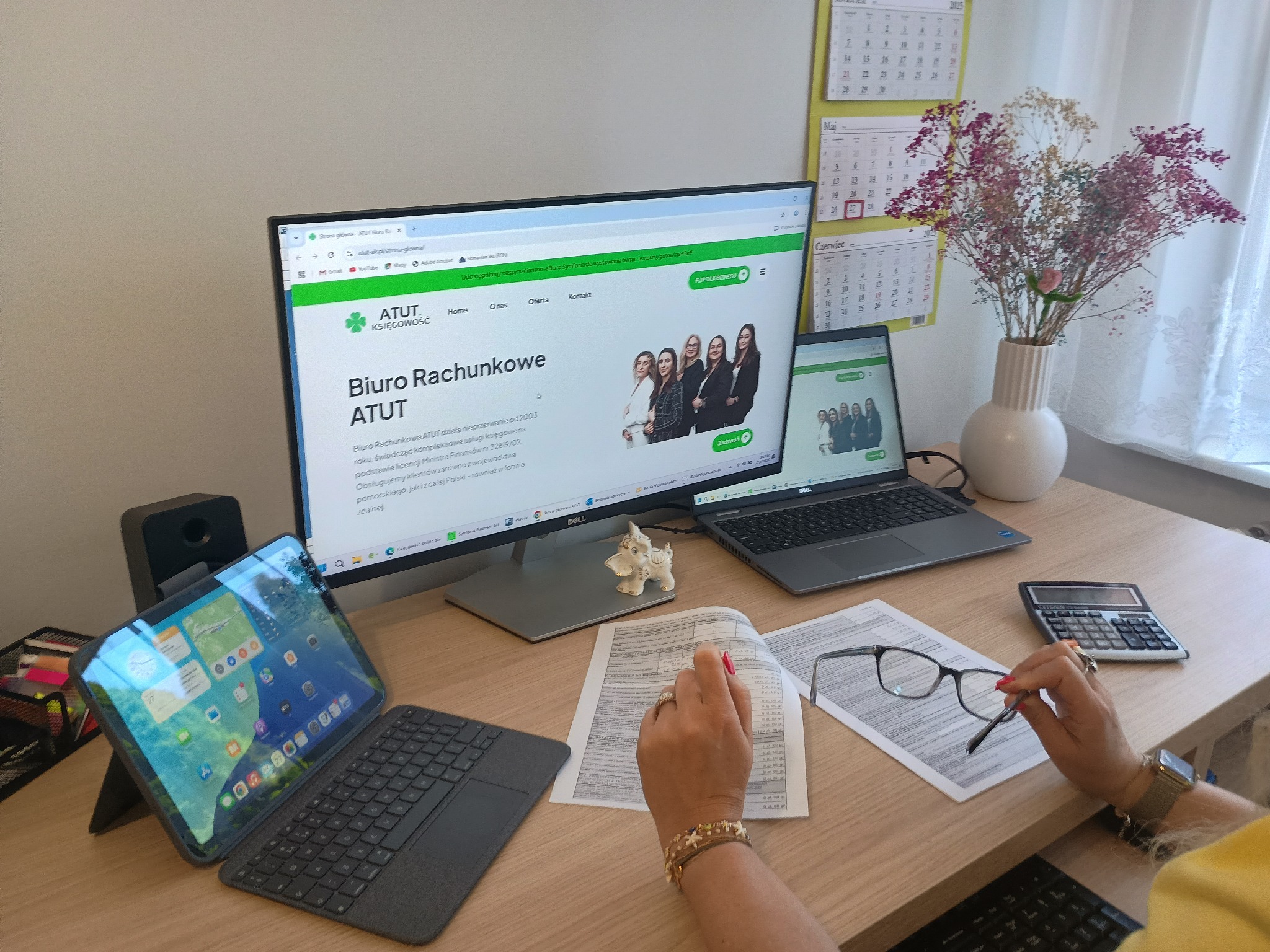 Biuro rachunkowe ATUT: monitor z otwartą stroną, laptop, tablet, dokumenty, kalkulator, okulary. Kobieta przegląda dokumenty na biurku.
