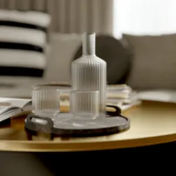 Stylowy stolik kawowy z czarną podstawą i złotym blatem. Na nim szklana karafka, dwa szklane naczynia na tacy i otwarta książka. W tle sofa z poduszkami.