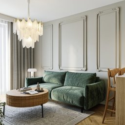 Stylowy salon z zieloną sofą, okrągłym stolikiem kawowym i designerską lampą. Neutralna kolorystyka ścian i podłóg. Uwagę przykuwają dekoracyjne ramy na ścianie.