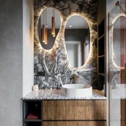 Niezabudka Design - Nowoczesna łazienka z umywalką nablatową, szafką z drewnianym fornirem i podświetlanym lustrem o nieregularnym kształcie na tle tapety w leśny motyw.