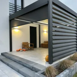 Nowoczesna, grafitowa pergola lamelowa z oświetleniem LED, przylegająca do jasnego budynku. Na tarasie drewniane meble i dekoracyjne dynie, dodające jesiennego klimatu.