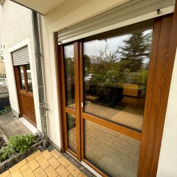 Okna, Rolety i Drzwi zewnętrzne - Pustułka - Żaluzje fasadowe i Pergole ogrodowe - Brązowe okno przesuwne z roletą zewnętrzną na tarasie z żółtej kostki. Widoczne odbicie wnętrza domu. Dwa okna z brązowymi ramami.