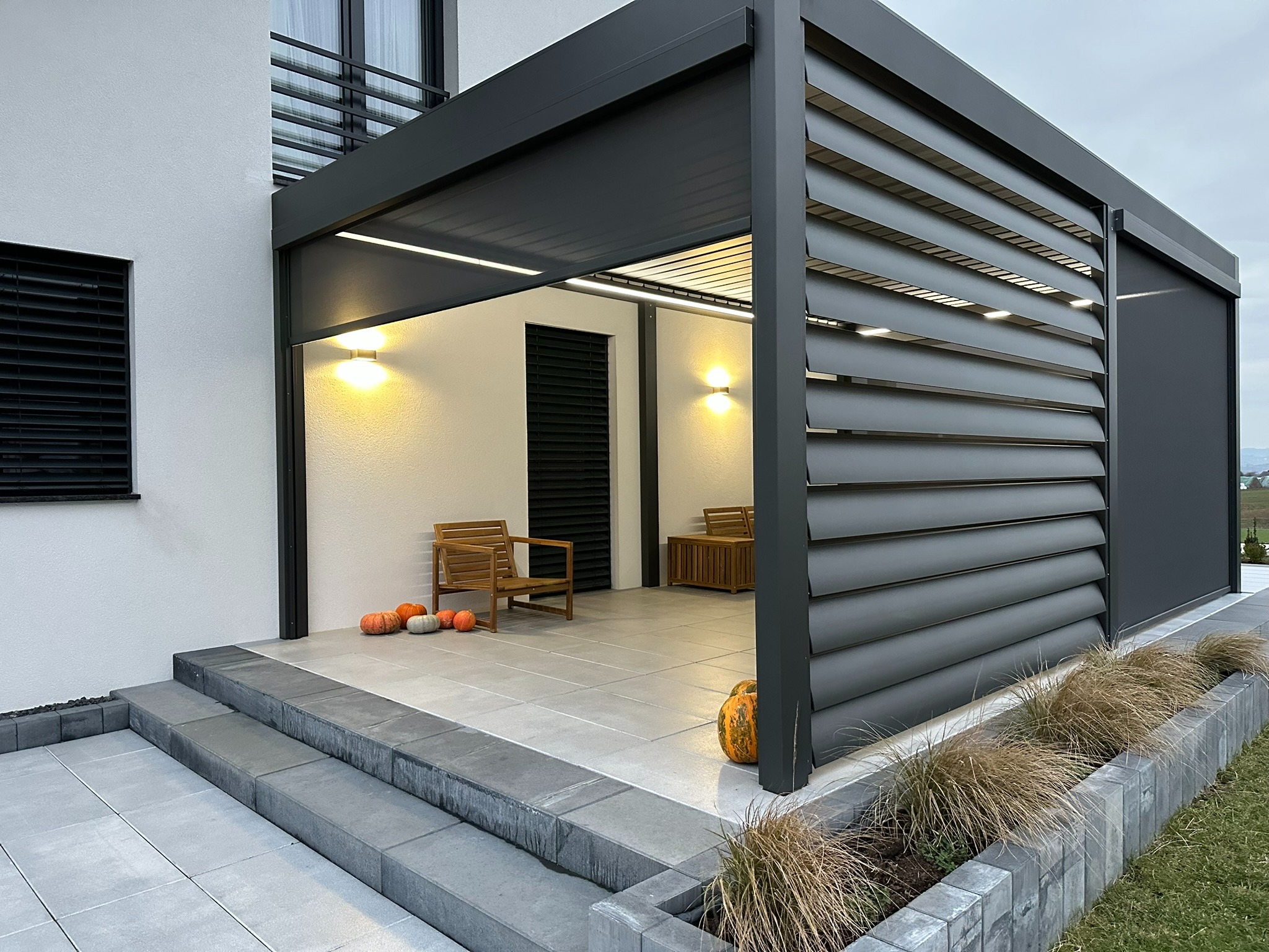 Nowoczesna, grafitowa pergola lamelowa z oświetleniem LED, przylegająca do jasnego budynku. Na tarasie drewniane meble i dekoracyjne dynie, dodające jesiennego klimatu.