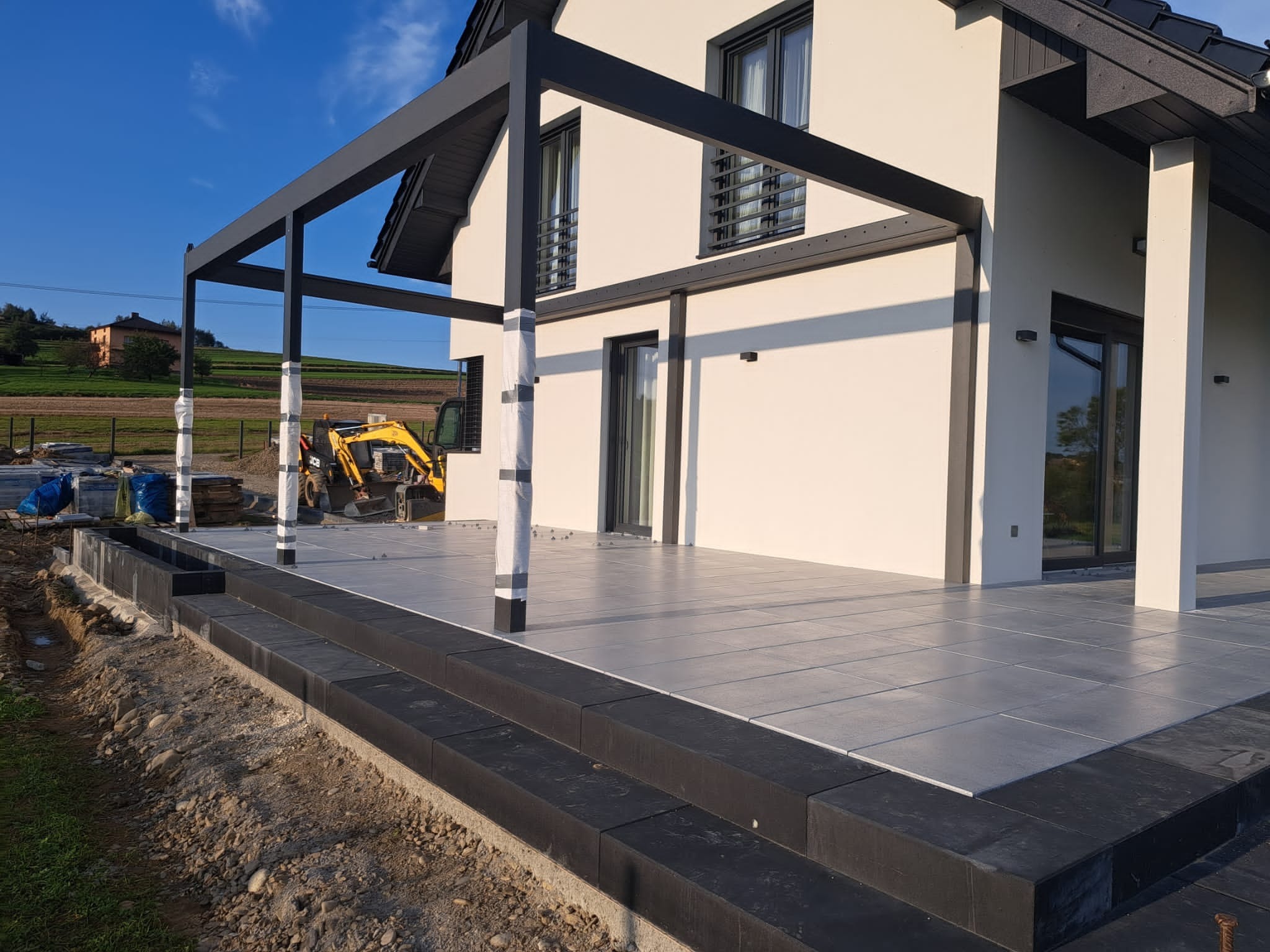 Nowoczesny taras z szarych płyt i czarną pergolą w trakcie budowy; w tle widoczny krajobraz i maszyna budowlana.