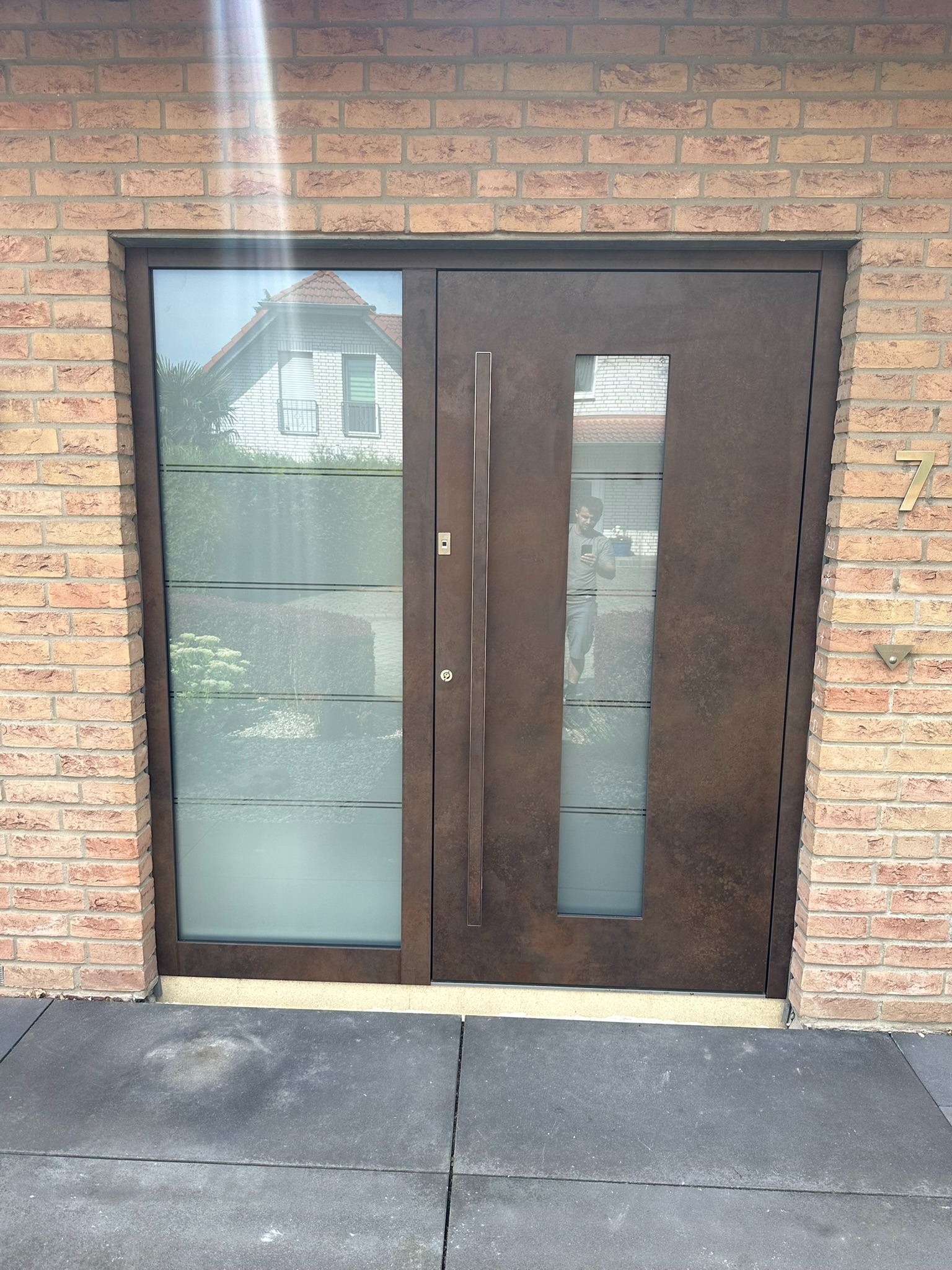 Nowoczesne, brązowe drzwi wejściowe z przeszkleniami, wkomponowane w elewację z cegły. Widoczne odbicie fotografa w szybie i numer domu '7' po prawej stronie.