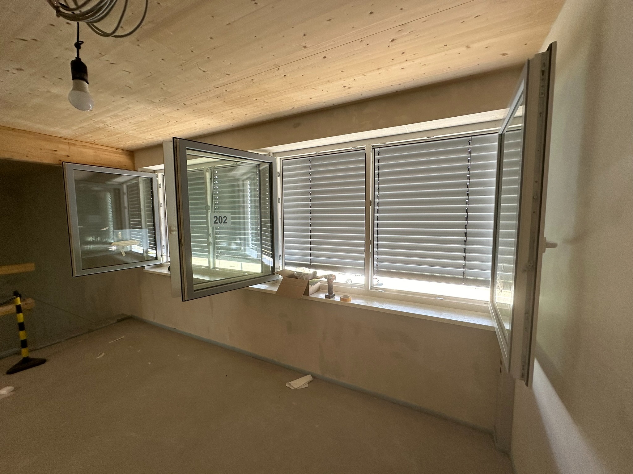Wnętrze z trzema oknami PCV, z roletami wewnętrznymi. Dwa okna uchylone, jedno zamknięte. Widoczne narzędzia i materiały montażowe na parapecie. Surowy stan wykończenia.