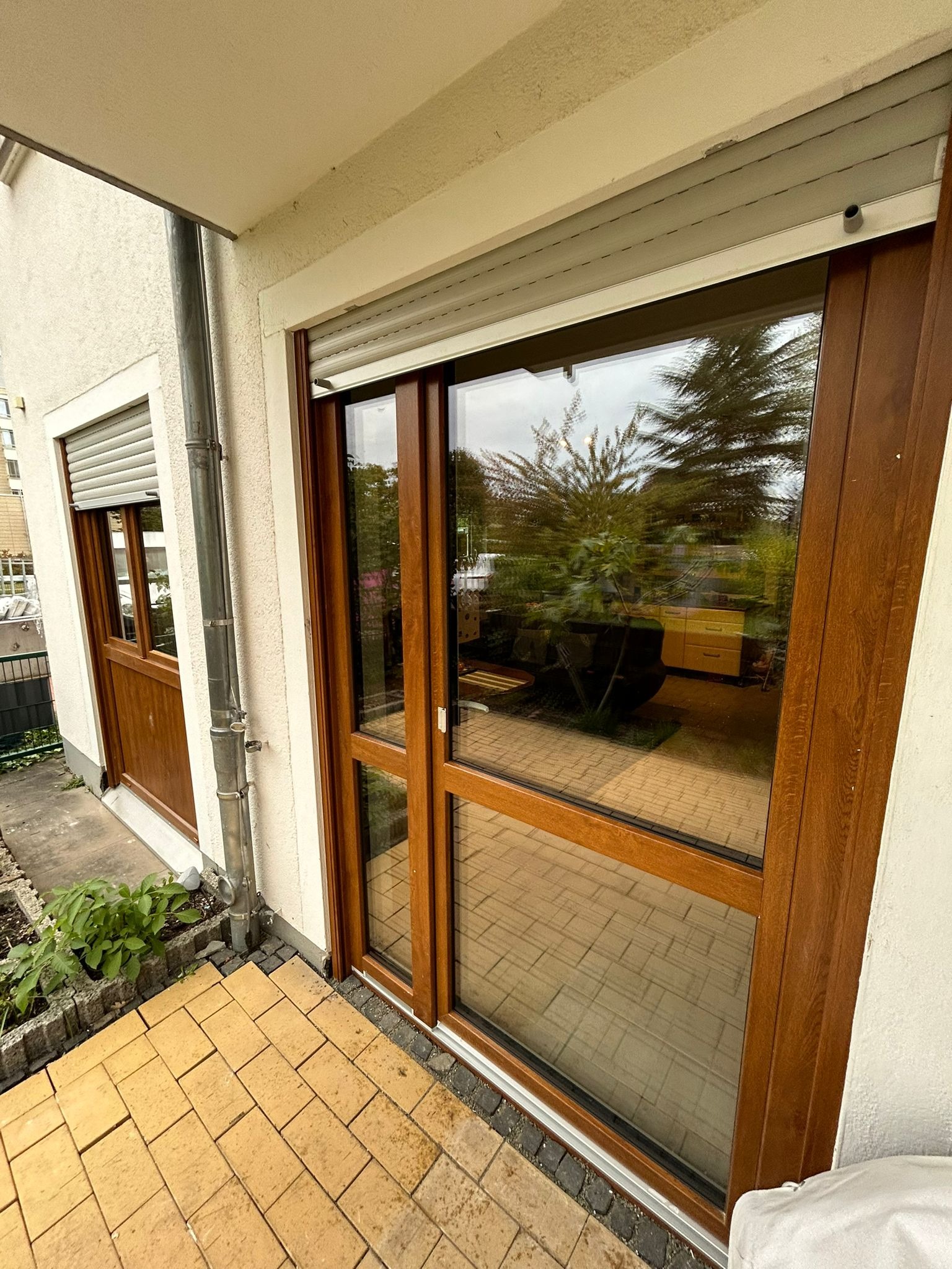 Brązowe okno przesuwne z roletą zewnętrzną na tarasie z żółtej kostki. Widoczne odbicie wnętrza domu. Dwa okna z brązowymi ramami.