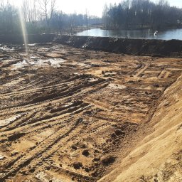 RTD Radosław Trzebiatowski - Wykopany teren pod zbiornik wodny z widocznymi śladami opon i brzegiem stawu w tle, oświetlony słonecznym światłem.