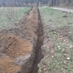 RTD Radosław Trzebiatowski - Wąski wykop w ziemi z ułożonym kablem, widok z góry, trawa i drzewa w tle, jesienna sceneria. Świeżo wykopana ziemia tworzy wały po obu stronach wykopu.