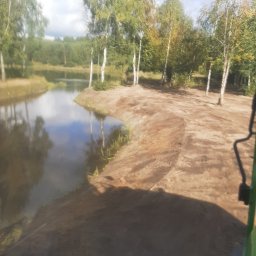 RTD Radosław Trzebiatowski - Krajobraz po wykopaniu stawu, z brzegami z piasku i odbiciem drzew w wodzie, widziany z perspektywy operatora maszyny. Błękitne niebo z chmurami w tle.