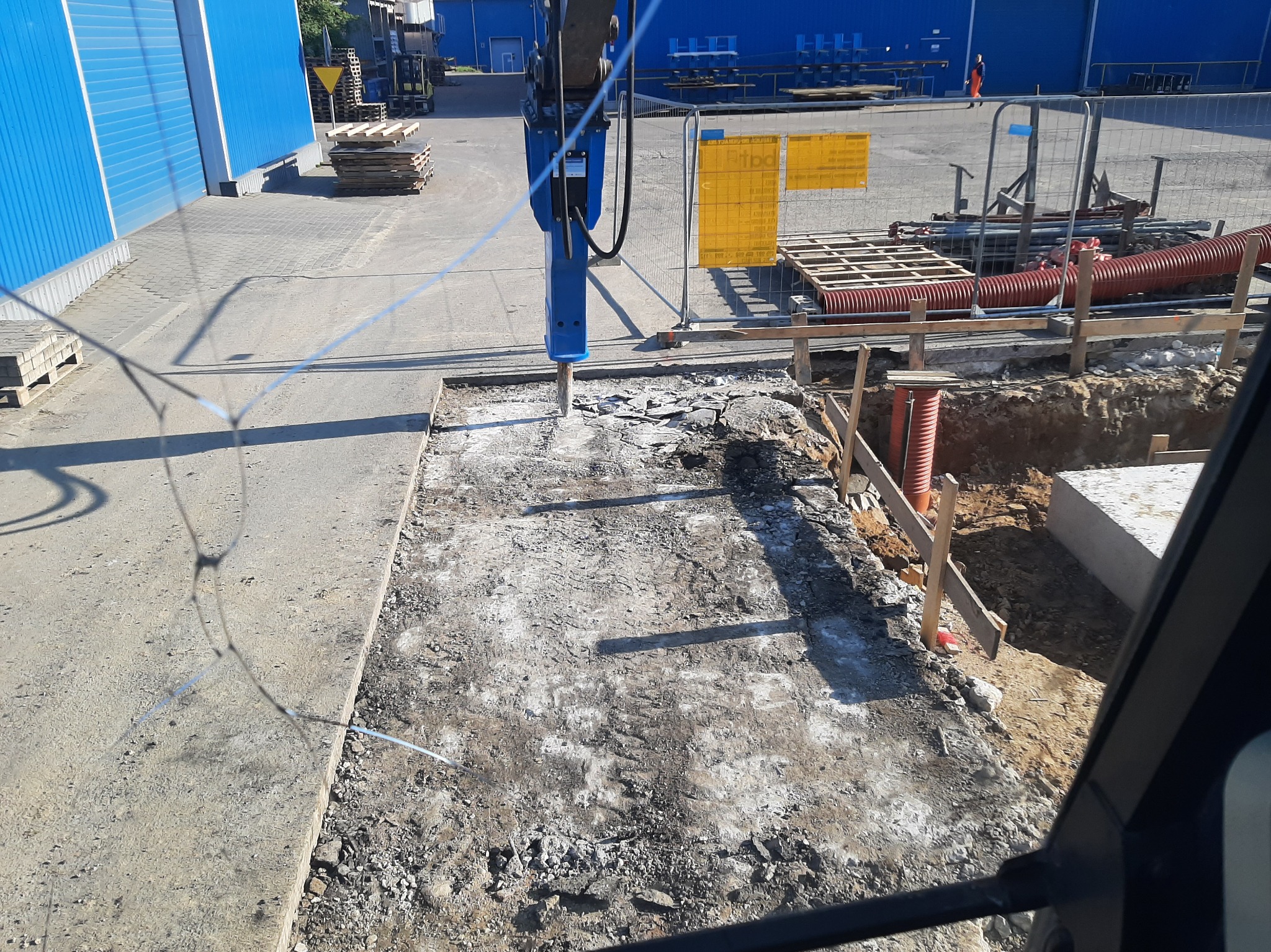 Niwelacja terenu pod budowę: Młot pneumatyczny kruszy betonową nawierzchnię. W tle widoczne materiały budowlane i fragment niebieskiej hali.