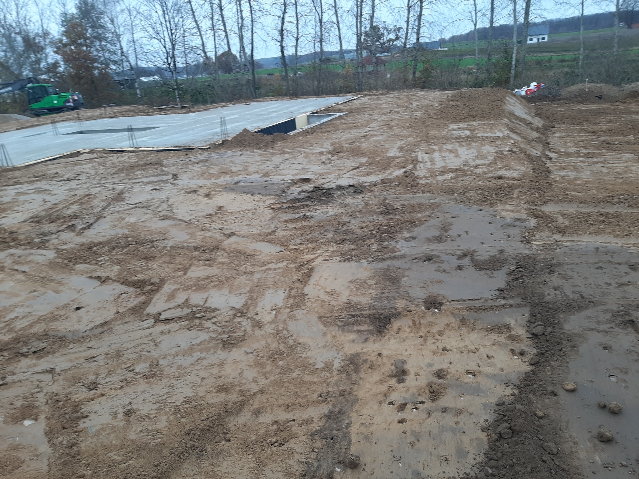 Wylany fundament z wystającymi prętami zbrojeniowymi, teren przygotowany pod budowę, w tle widoczny zielony sprzęt budowlany i drzewa.