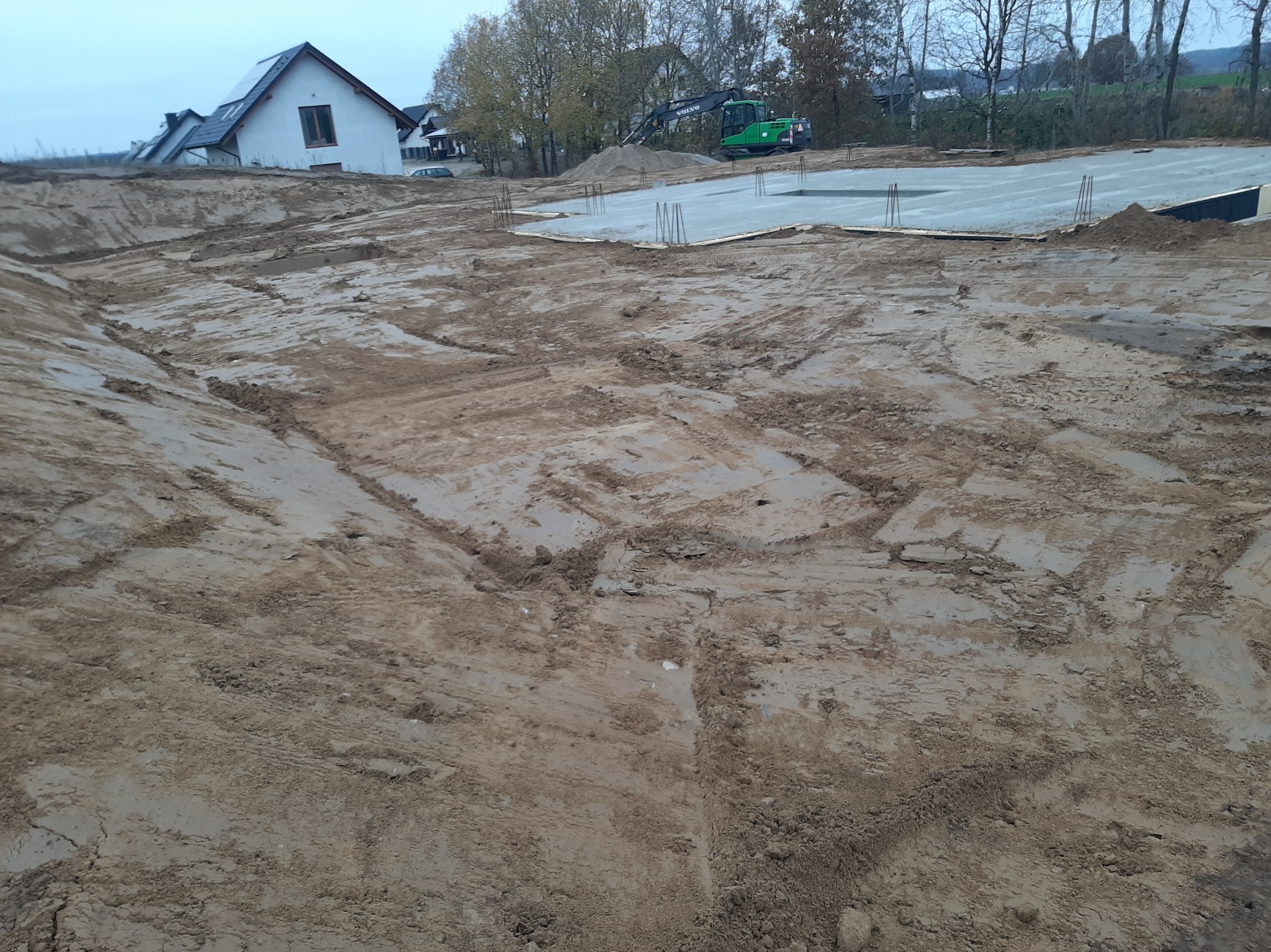 Wyrównany teren pod budowę domu z wylanym fundamentem i wystającymi prętami zbrojeniowymi. W tle zielona koparka Volvo i biały dom z szarym dachem.