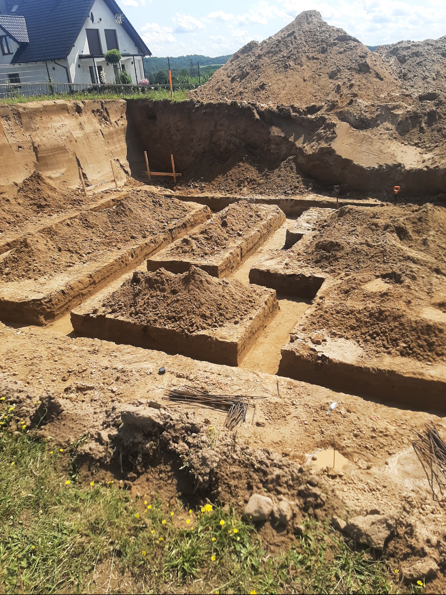 Wykopane rowy pod fundamenty domu jednorodzinnego z widoczną stertą ziemi i fragmentem budynku w tle, wykonane w słoneczny dzień.