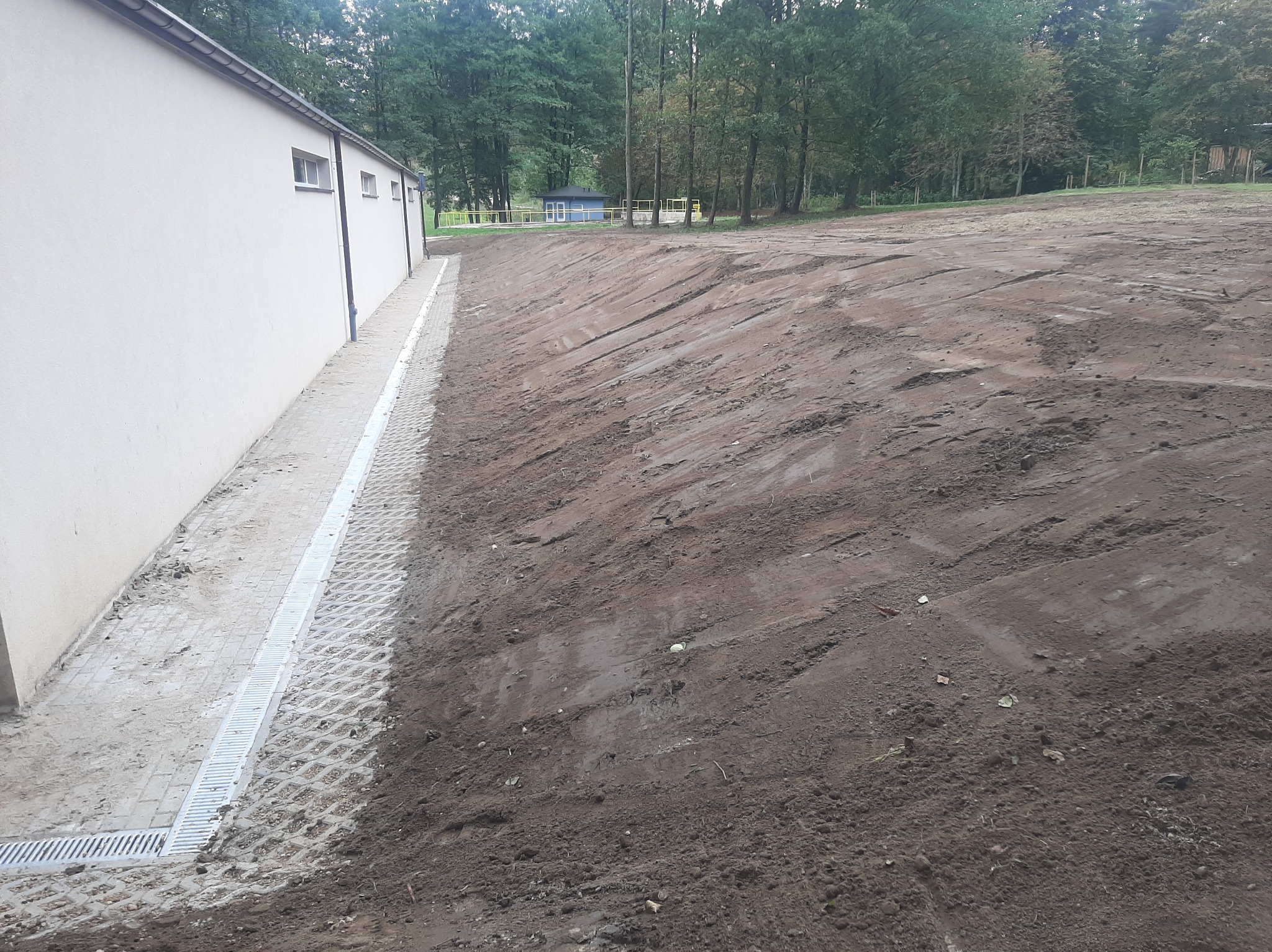 Ukształtowany teren z drenażem przy budynku. Widoczne ślady maszyn, przygotowany grunt pod dalsze prace. W tle zieleń drzew.