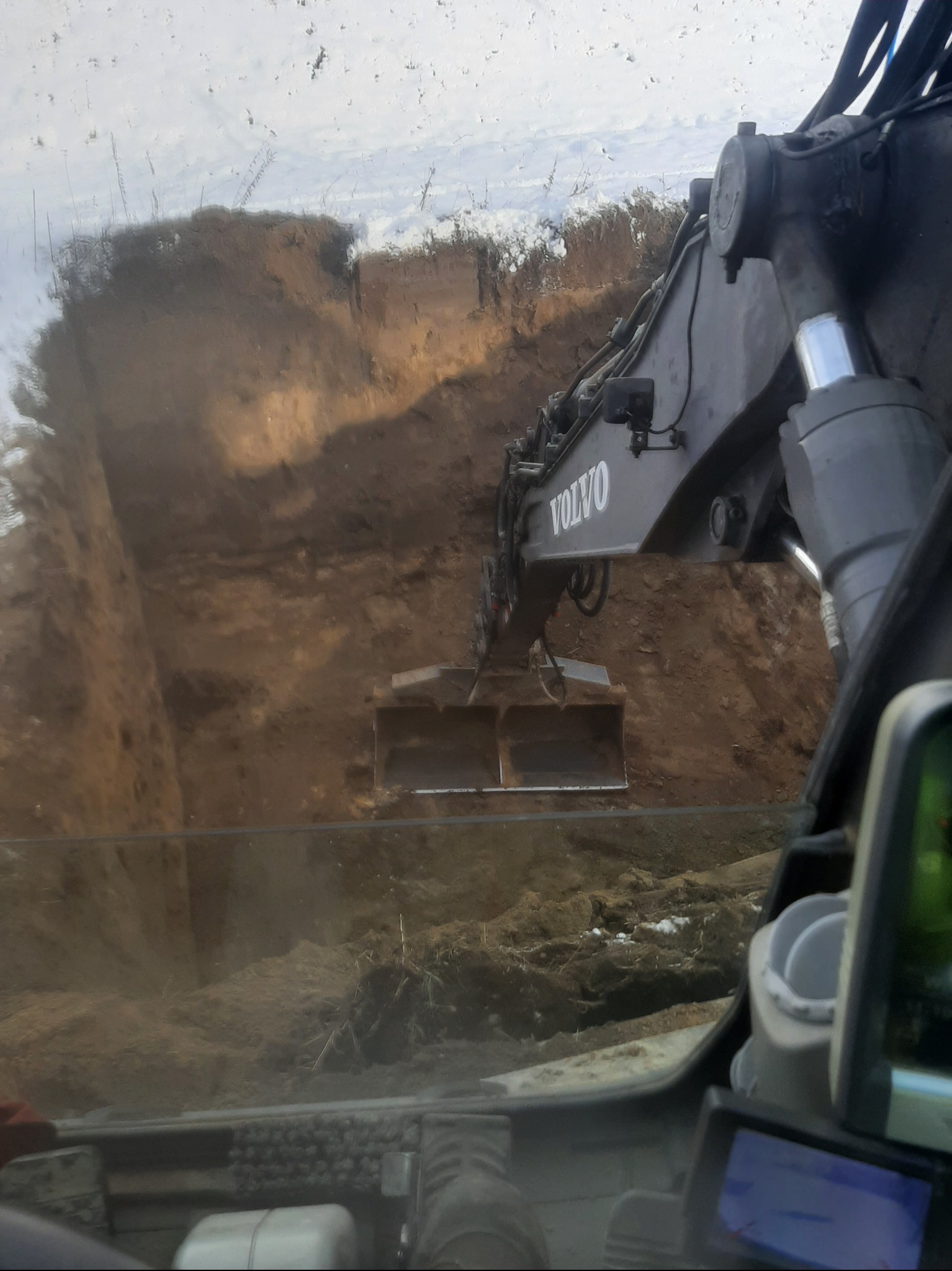 Koparka Volvo w trakcie wykopu ziemi, widok z kabiny operatora, ziemia częściowo pokryta śniegiem. Prace ziemne w trakcie realizacji.