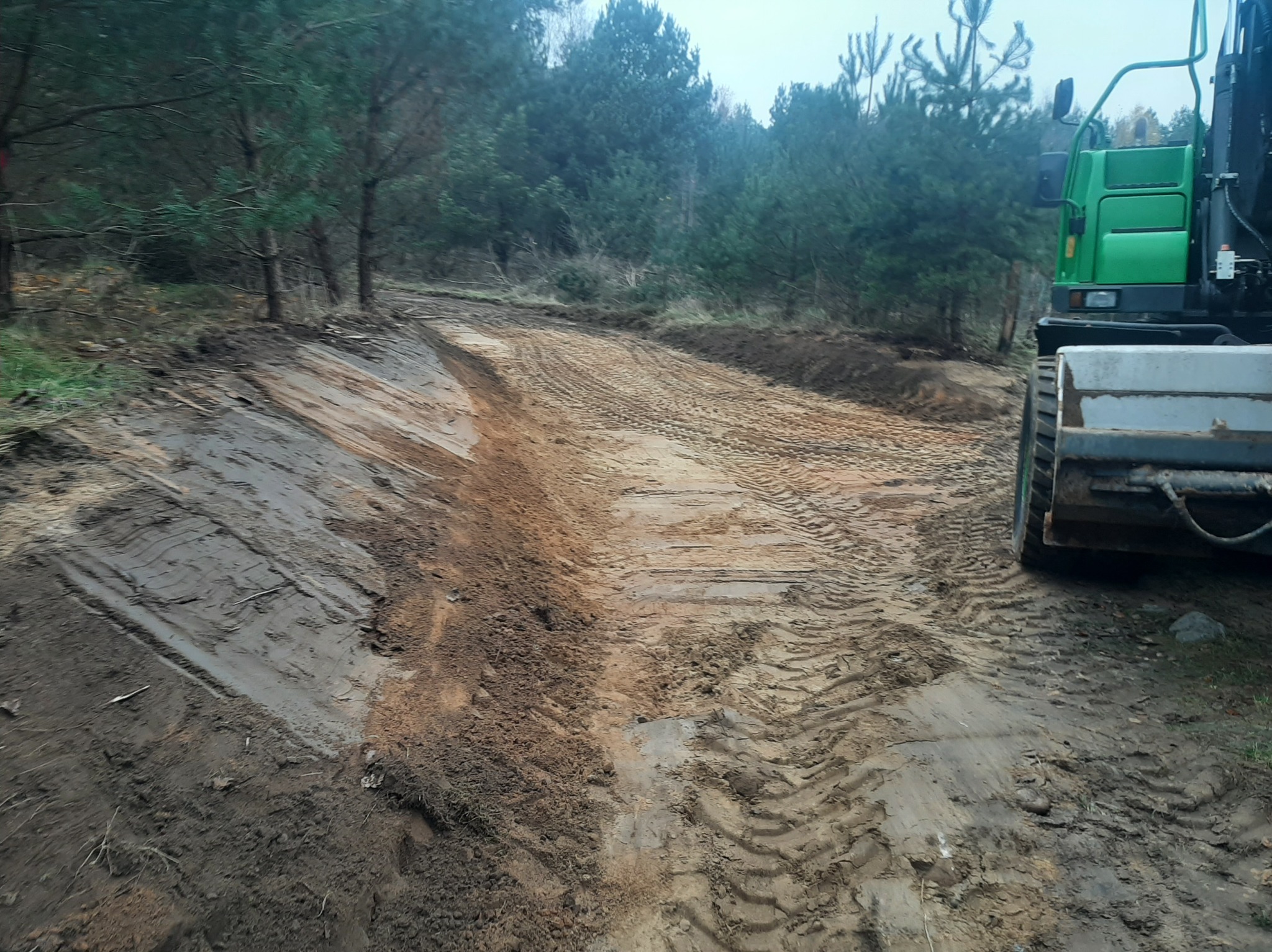 Niwelacja terenu leśnego z użyciem zielonej maszyny budowlanej. Ślady opon na ubitej ziemi. Prace ziemne w toku, widoczne wyprofilowane zbocza.