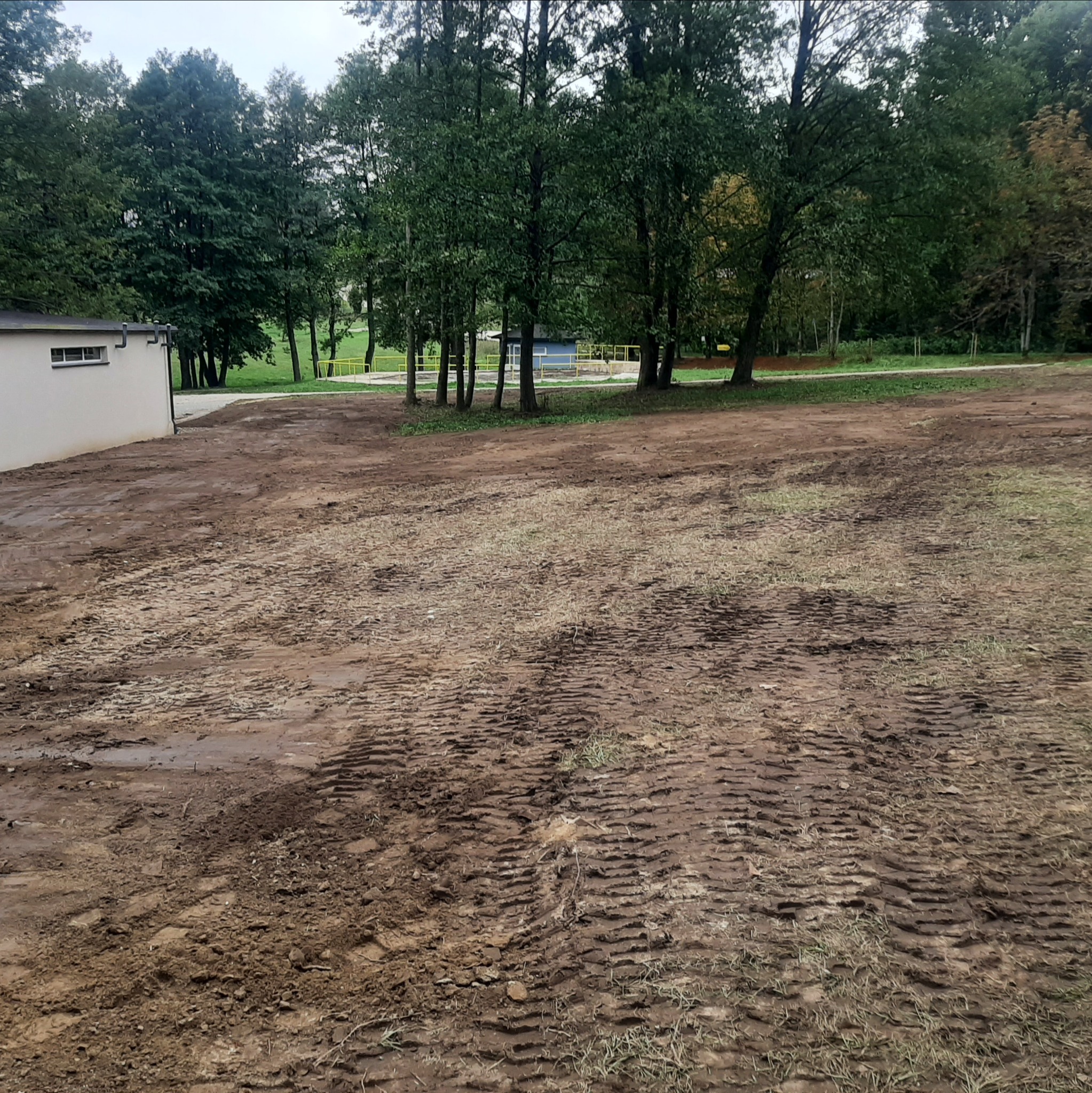 Ujęcie z poziomu gruntu na wyrównany, wilgotny teren z widocznymi śladami opon, otoczony drzewami i budynkiem w tle. Krajobraz po niwelacji.