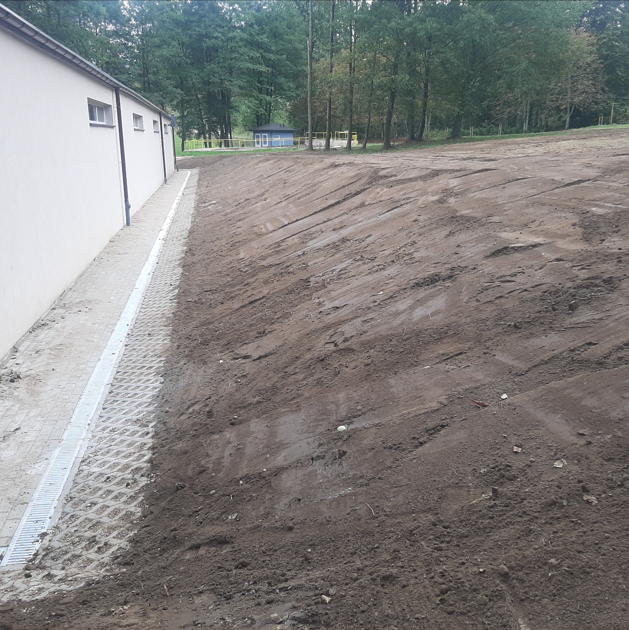 Ukończona niwelacja terenu obok budynku z drenażem, widoczne ślady po maszynach. W tle drzewa i mały budynek.