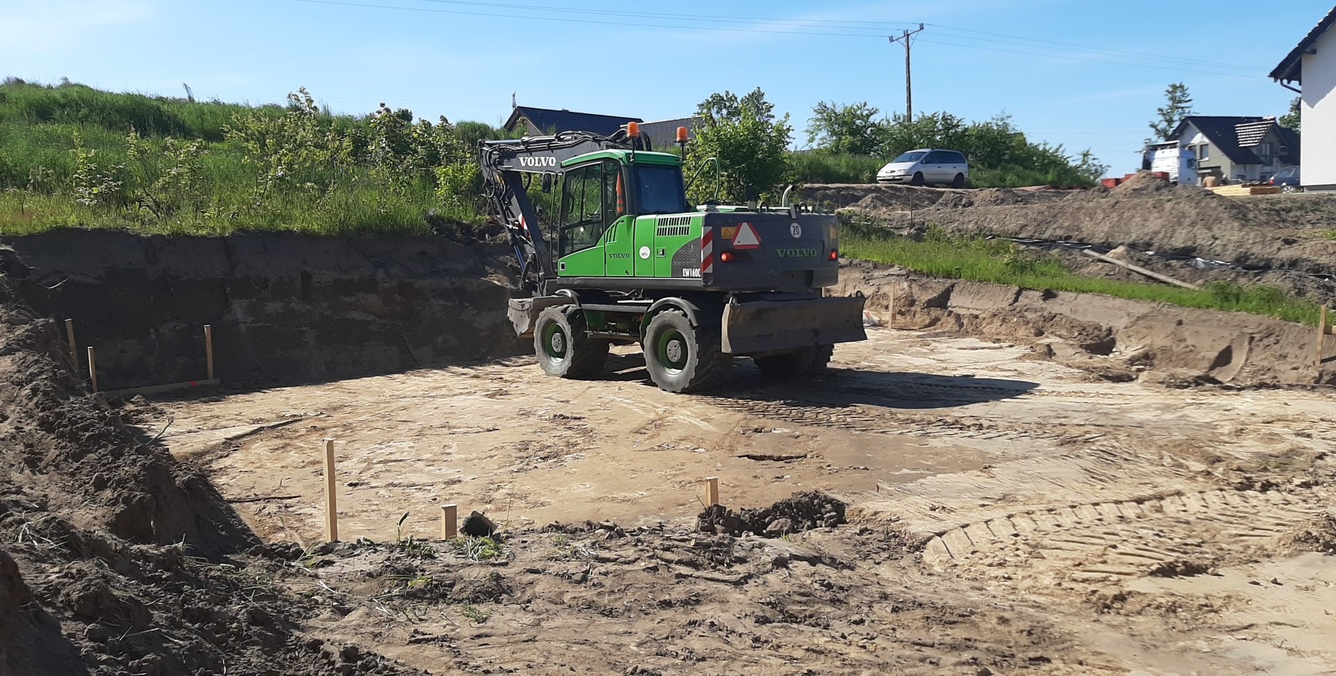 Zielona koparka Volvo EW160C na placu budowy, wykop pod fundamenty z wbitymi drewnianymi palikami, w tle zabudowa jednorodzinna i błękitne niebo.