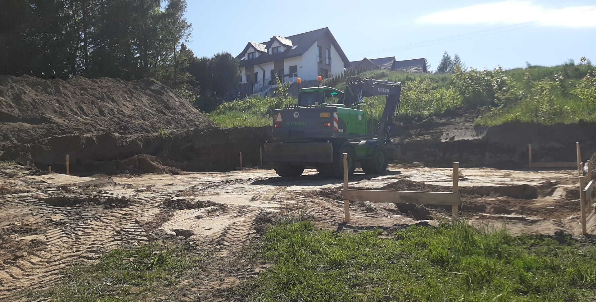 Zielona koparka Volvo na wykopie pod fundamenty domu jednorodzinnego, widoczne drewniane szalunki i hałda ziemi. Słoneczny dzień.