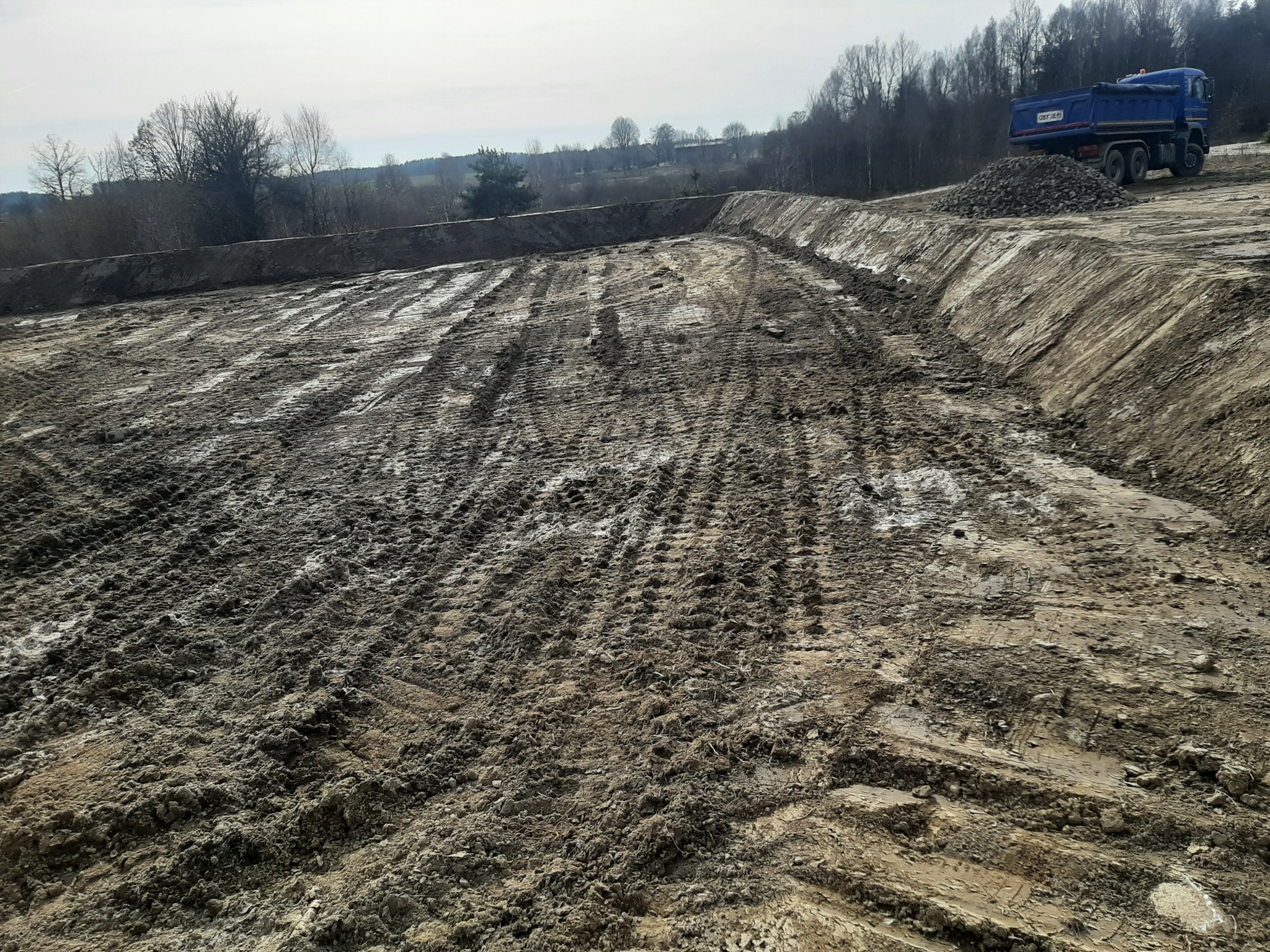 Wykopany teren z widocznymi śladami opon i stertą gruzu w Bytowie. W tle drzewa i niebieska ciężarówka. Prace ziemne w toku.