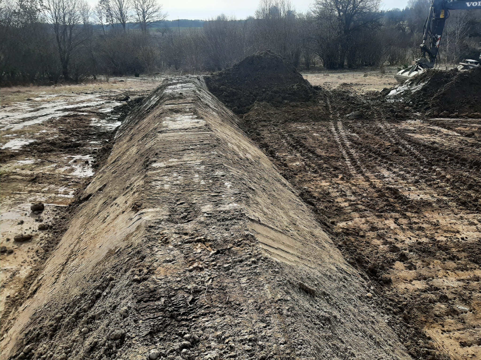 Wyrównywanie terenu z użyciem koparki, widoczne ślady opon i świeżo usypana ziemia, w tle drzewa i zarys pól. Prace ziemne w toku.
