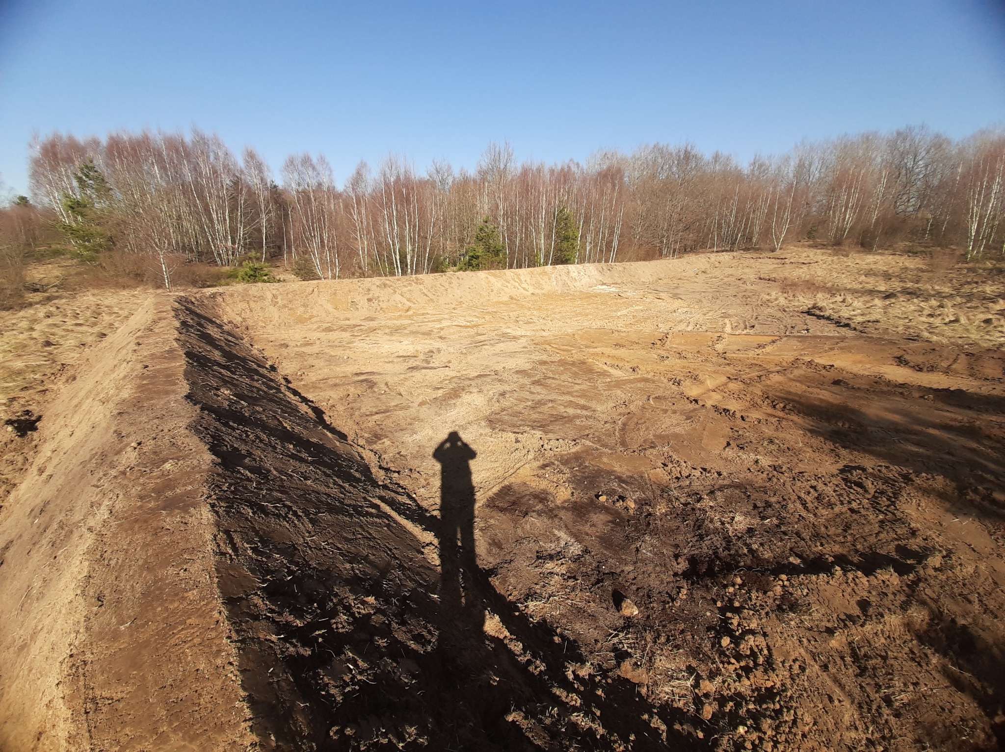 Wyrobisko ziemne z shadow selfie na pierwszym planie, w tle drzewa bez liści i błękitne niebo. Teren przygotowany pod inwestycję.