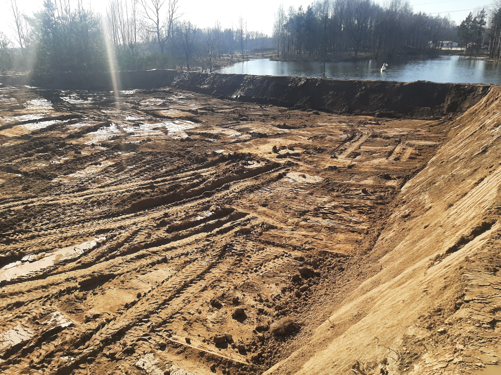 Wykopany teren pod zbiornik wodny z widocznymi śladami opon i brzegiem stawu w tle, oświetlony słonecznym światłem.