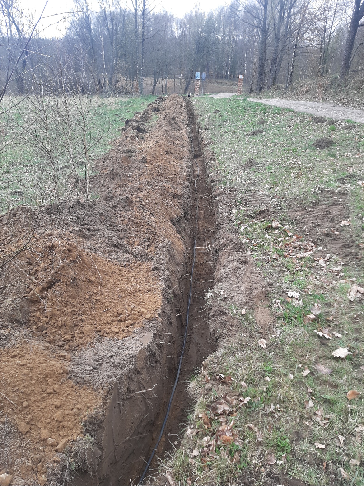 Wąski wykop w ziemi z ułożonym kablem, widok z góry, trawa i drzewa w tle, jesienna sceneria. Świeżo wykopana ziemia tworzy wały po obu stronach wykopu.