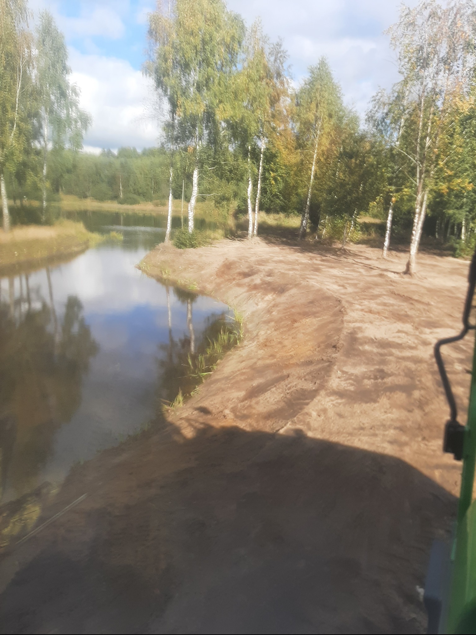 Krajobraz po wykopaniu stawu, z brzegami z piasku i odbiciem drzew w wodzie, widziany z perspektywy operatora maszyny. Błękitne niebo z chmurami w tle.