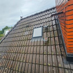 Washingservice - Mokra, brudna dachówka z mchem i małym oknem dachowym, przylegająca do ceglanego komina z metalowym wspornikiem na tle pochmurnego nieba.