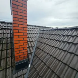 Ceglany komin z ciemnymi fugami i mokry, ciemnobrązowy dach pokryty dachówką karpiówką pod pochmurnym niebem. Widok z góry.