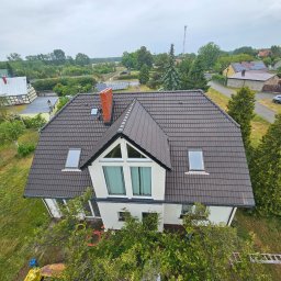 Washingservice - Dom jednorodzinny z brązowym dachem z dachówki, widok z góry. Biała fasada, okna dachowe i komin z czerwonej cegły. W tle zieleń drzew i zabudowa mieszkalna.