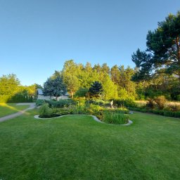 DEGO Group Sp. z o.o. - Rozległy, zielony trawnik z oczkiem wodnym i różnorodną roślinnością. W tle drzewa iglaste i liściaste, dom i osoba spacerująca po ogrodzie w słoneczny dzień.