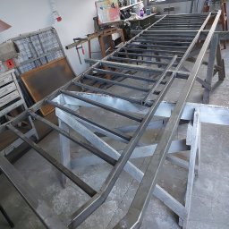 DEGO Group Sp. z o.o. - Spawana konstrukcja stalowa, prawdopodobnie rama, na drewnianych kozłach w warsztacie. Widoczne ślady spawania i obróbki metalu. W tle narzędzia i wyposażenie warsztatowe.