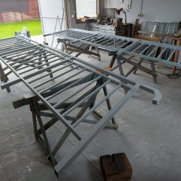 DEGO Group Sp. z o.o. - Dwie pomalowane na szaro metalowe konstrukcje na drewnianych kozłach w warsztacie. W tle narzędzia i fragment ogrodu. Widok z góry, lekko z ukosa.