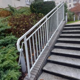 DEGO Group Sp. z o.o. - Schody z jasnego kamienia z białą barierką, otoczone zielenią krzewów. Jesienne liście na stopniach. Budynek mieszkalny w tle, pochmurne niebo.