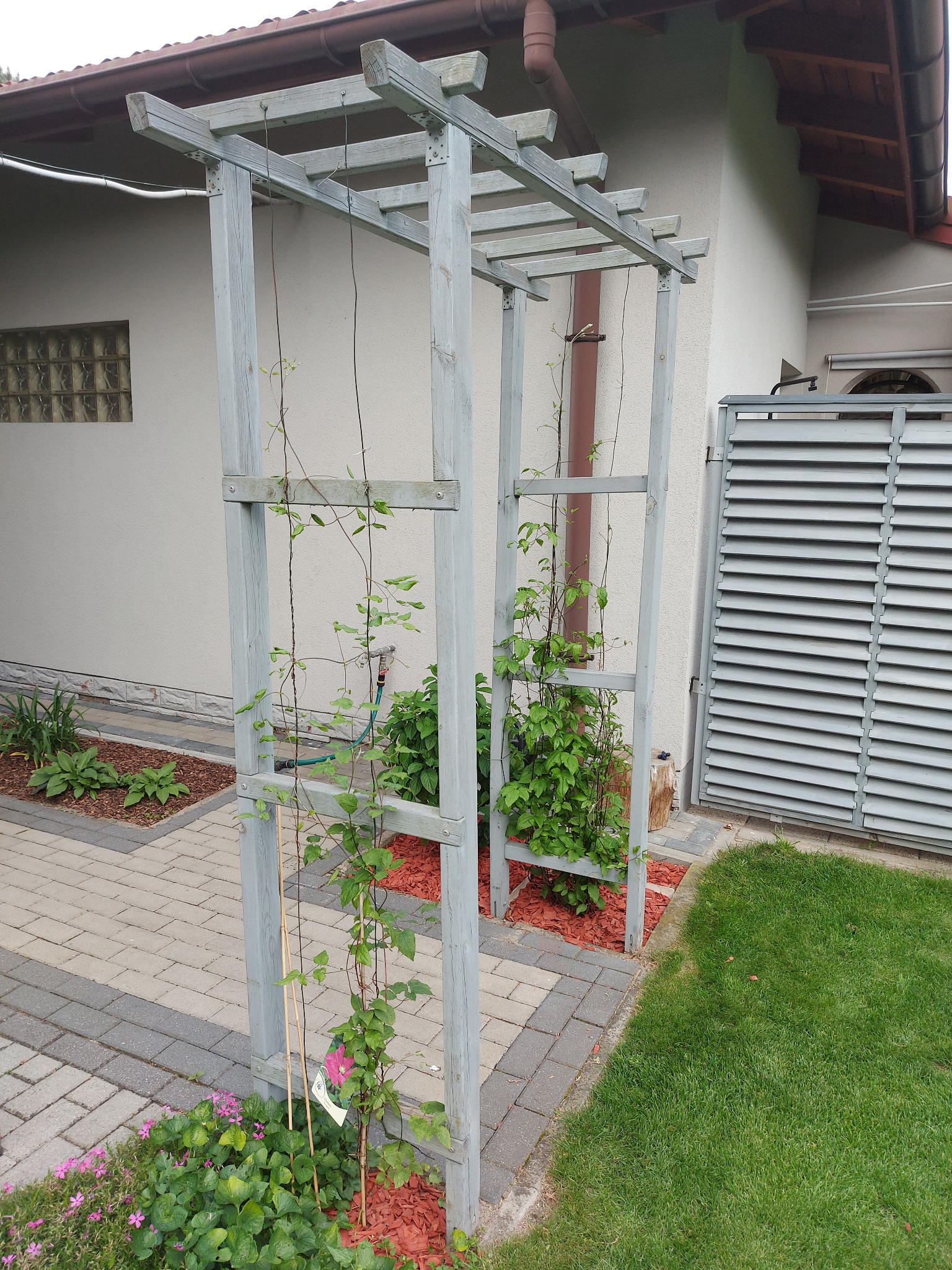 Drewniana pergola z pnącymi roślinami w ogrodzie, obok ścieżki z kostki i trawnika. Widoczny fragment elewacji budynku z osłoną wentylacyjną.