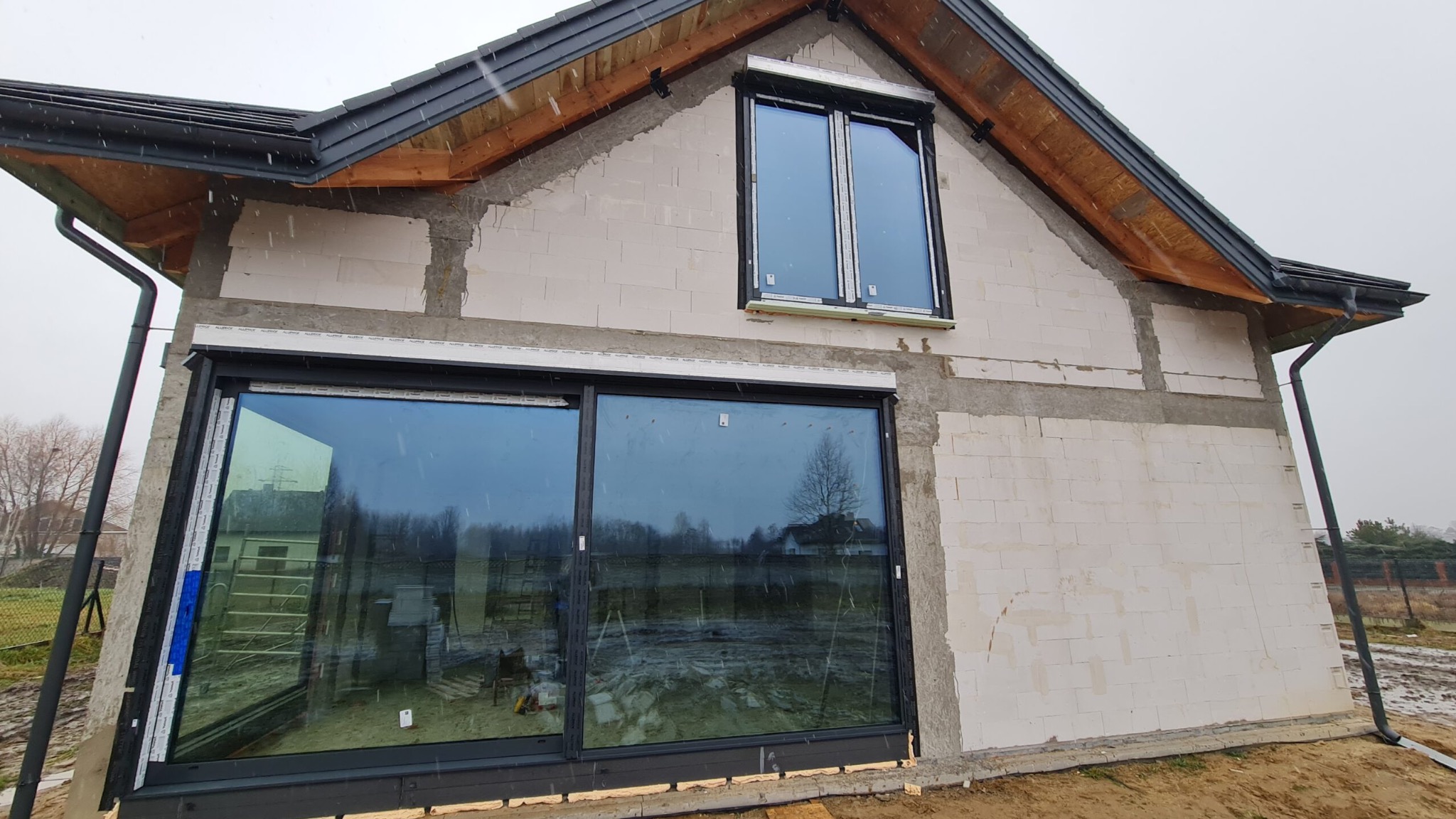 Dom w trakcie budowy z zamontowanymi oknami przesuwnymi i mniejszym oknem na górze. Widoczne bloczki betonowe i niedokończona elewacja. Szare, pochmurne niebo.