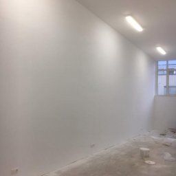 Malta - Wnętrze po malowaniu: gładkie, białe ściany, surowa podłoga, widoczne gniazdka. Minimalistyczny efekt odświeżenia z widokiem na okno w tle.