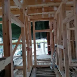 Wnętrze domu w trakcie budowy, widoczna drewniana konstrukcja szkieletowa, schody betonowe i drabina oparta o ścianę. Surowy stan, widoczne elementy instalacji.